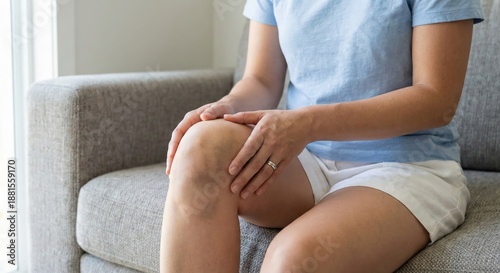 A woman's hands gently examine a large purple bruise on her knee while sitting on a grey couch indoors, focusing on the injury and pain