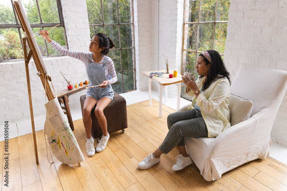 Fototapeta premium Asian teenage daughter and mother painting at easel in art studio with daughter holding palette