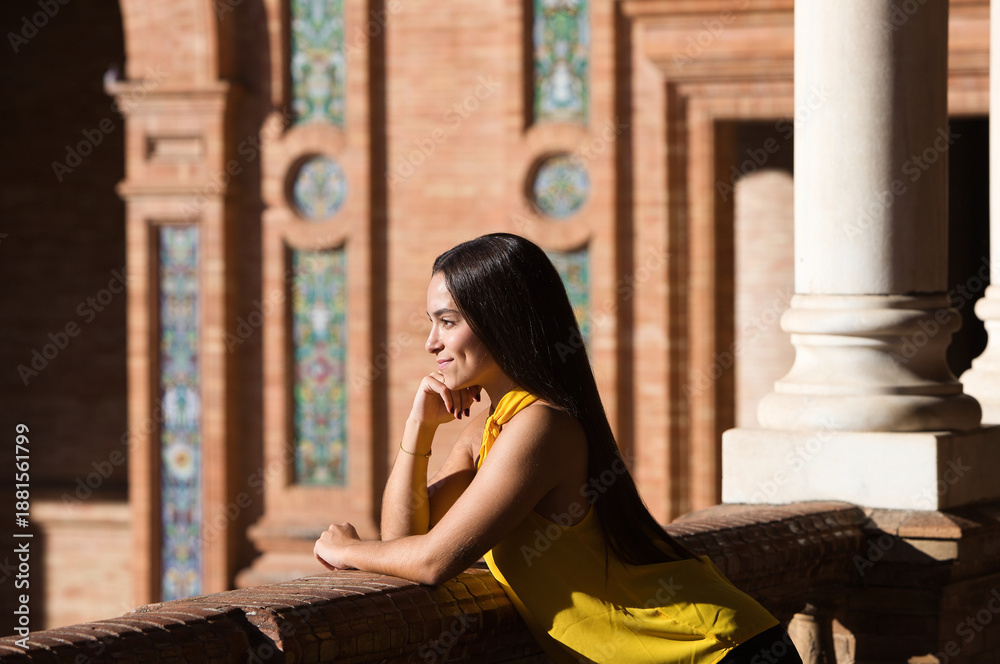 Fototapeta premium A young, beautiful, dark-haired Latin woman wearing a yellow shirt and black trousers leans on the railing of the Plaza de España with her chin resting on her hand, looking attentive and happy.