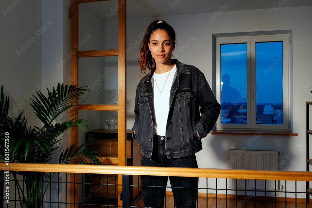 Fototapeta premium Portrait of young adult biracial woman standing indoors with hands in pockets looking into camera, confident expression, visible window and plant in background evening setting