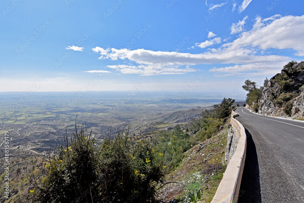Obraz premium Scenic mountain road curving alongside a steep cliff, overlooking a vast valley under a blue sky with soft white clouds. A wide, bright summer vista.