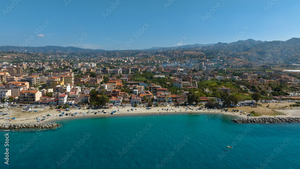 Fototapeta premium Aerial view of Gallico Beach, Calabria, Italy. It is a Mediterranean coastal town. Residential buildings line the sandy beach and turquoise sea, with lush green mountains rising in the distance.