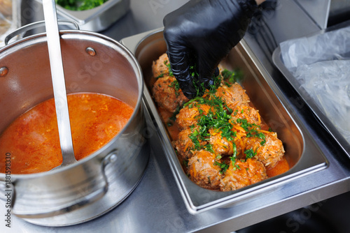 Chef hand with black glove garnishing cooked meatballs in a serving tray with fresh parsley and dill