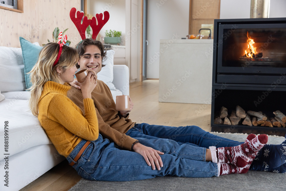 Fototapeta premium Couple sitting on rug in living room near fireplace wearing antler headbands holding ceramic mugs