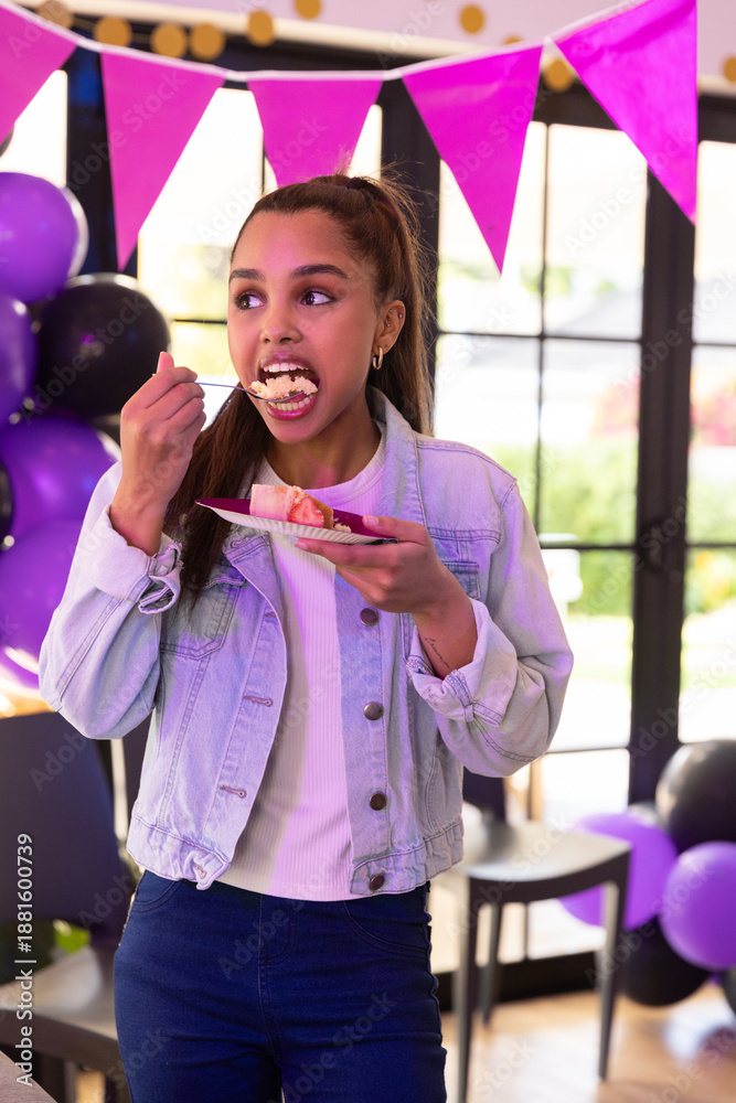 Obraz premium Adolescent girl standing and eating slice of cake from plate at party with balloons, banners