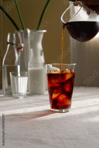 Fényképezés Cold brew coffee is being poured from a glass jar into a glass with ice on table covered with a natural linen tablecloth