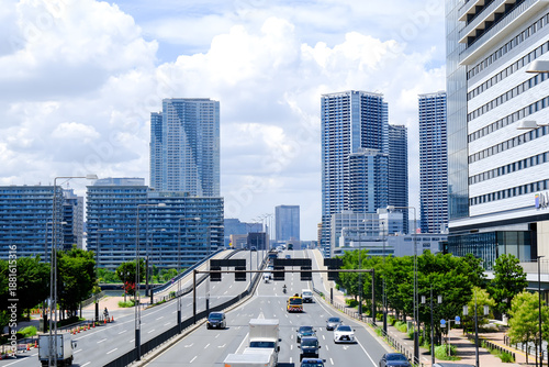 A wide-angle landscape photograph shows the skyline of a modern city along a river on a sunny day. Several tall skyscrapers with glass and concrete walls stand out in the right and center of Tokyo.