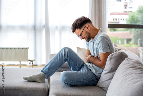Foto Young man sitting on sofa suffering from severe stomach ache and abdominal pain