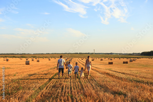 A family walks hand in hand through a golden wheat field during sunset. The scene captures a warm, peaceful atmosphere with hay bales scattered around.