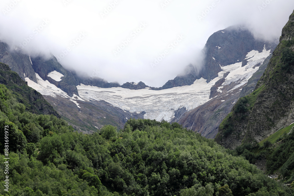 Fototapeta premium Green forest below mountain glacier with clouds