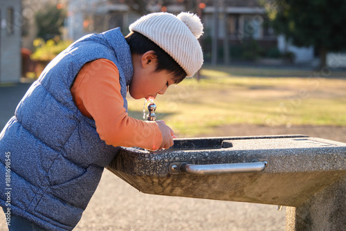 Tableau sur toile Japanese boy hydrating from a water fountain in a park wearing winter clothing