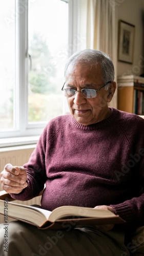 Vertical video of a senior man reading a book in a living room. Elderly person putting on glasses while sitting in an armchair. Retirement lifestyle and leisure concept