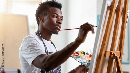 Focused African American male artist painting on canvas with brush and palette in creative studio.