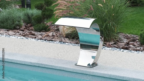 Modern stainless steel pool waterfall installed on a swimming pool edge, with clear blue water and landscaped garden background, conveying luxury, wellness, relaxation and premium outdoor design
