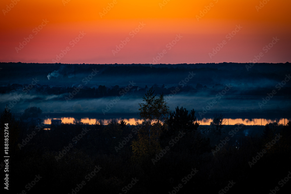 Obraz premium A serene sunset landscape with warm orange sky reflecting on a calm river, layered with misty fog over a rural village and forest horizon, creating a peaceful and atmospheric evening scene.