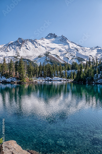 A mountain range is reflected in the water of a lake. The sky is clear and blue, and the snow on the mountains is white. The scene is peaceful and serene, with the water
