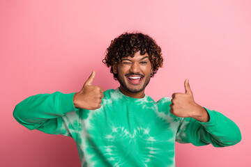 Young man with curly hair wearing green tie dye sweater gives thumbs up smiles at camera pink background for a joyful lifestyle advertising image