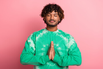 Young man with curly hair wearing a green tie dye sweater meditates with hands pressed together in front of a pink background