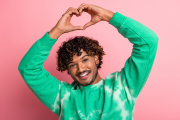 Young man with curly hair in a green tie dye sweater makes a heart shape with his arms against a pink background smiling warmly conveying joy friendship and fashionable lifestyle for advertising 