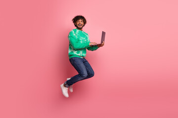 Young man jumps with laptop against pink background wearing green tie dye sweater and jeans