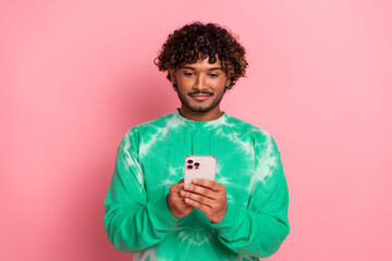 Young man with curly hair wearing a green tie dye sweater looks at his phone against pink background in a casual lifestyle shot