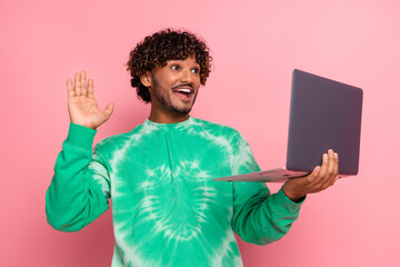 Young man with curly hair wearing a green tie dye sweater holds a laptop and waves with a big smile in a pink studio