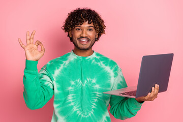 Young male model in green tie dye sweater holds laptop and smiles against pink background