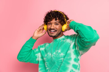 Young man with yellow headphones wearing a green tie dye sweater smiles against a pink background in a casual lifestyle photo shoot
