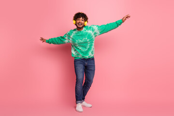 Young man with curly hair wearing a green tie dye sweater dancing against pink background with headphones