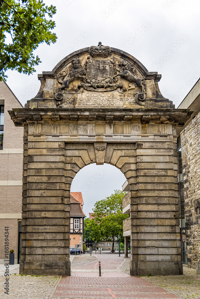 Obraz premium A stone archway in Hanover Germany with a crown on top of it. The archway is in front of a building with a red roof