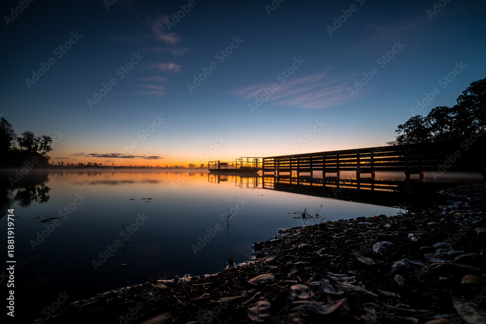 Fototapeta premium A beautiful sunset over a lake with a wooden bridge in the foreground. The water is calm and the sky is filled with clouds. The scene is peaceful and serene