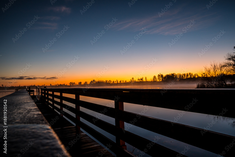 Obraz premium A wooden bridge over a body of water with a beautiful sunset in the background. The water is calm and the sky is filled with clouds