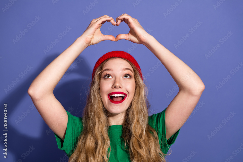 Fototapeta premium Happy young woman forms a heart with her hands against a purple background wear red beanie and green shirt in a playful fashionable photo