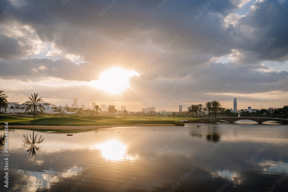 Obraz premium Dubai, United Arab Emirates - January 17, 2026: Sunset over city skyline reflecting in calm water with palm trees and bridge in foreground