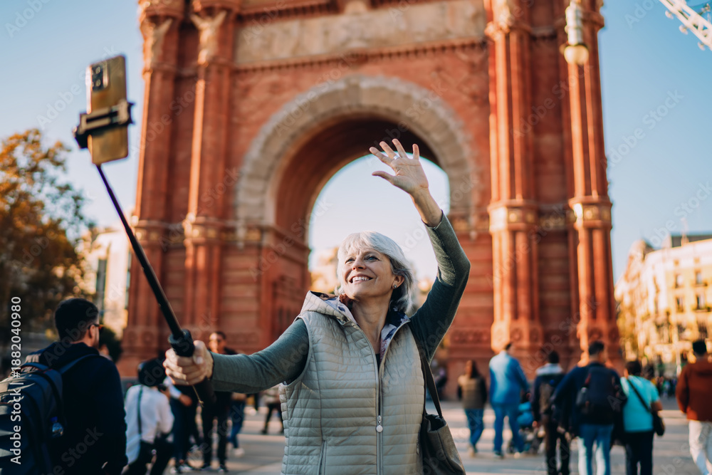 Obraz premium Older woman waves cheerfully while filming selfie video with smartphone and selfie stick. A lively scene showing age diversity and natural confidence with mobile tech.