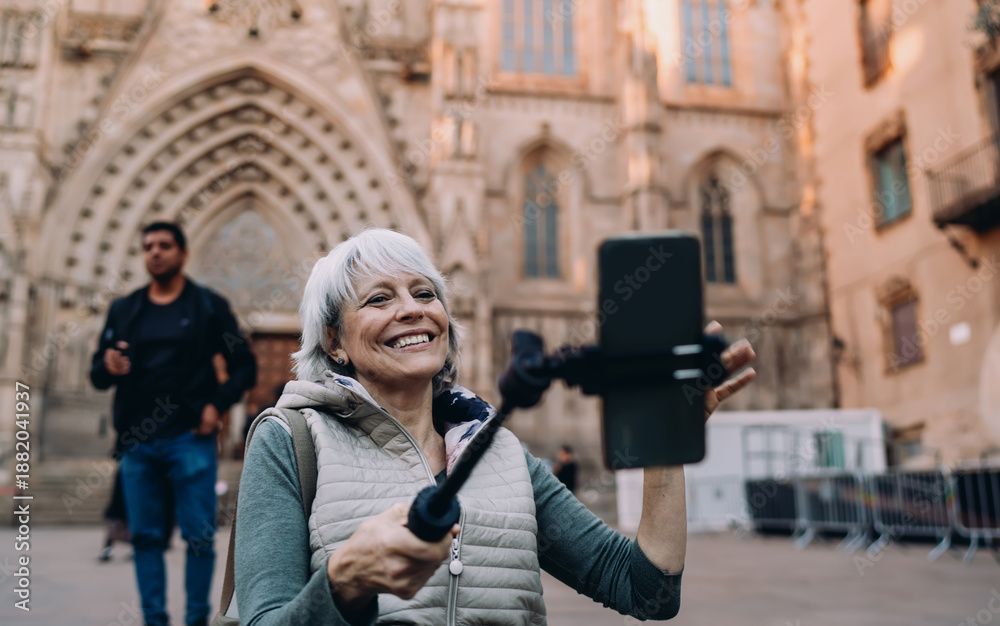 Fototapeta premium Happy senior woman recording herself on smartphone with selfie stick in front of historical cathedral. Captures joyful solo travel, digital expression and confident aging.