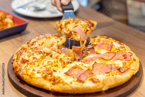 A freshly Melted cheese stretches baked pizza sits on a wooden board as a slice is being lifted by hand . Ham and pineapple rest over bubbling cheese on a round pizza on wooden table 