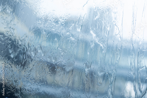 A rainy water droplets run down a wet glass reflects light surface while water trails move downward , blurring the view behind it.