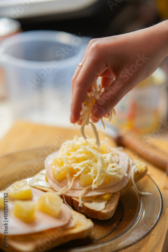 Hand adding shredded cheese to open-faced Hawaiian toasts with ham and pineapple for a delicious meal.