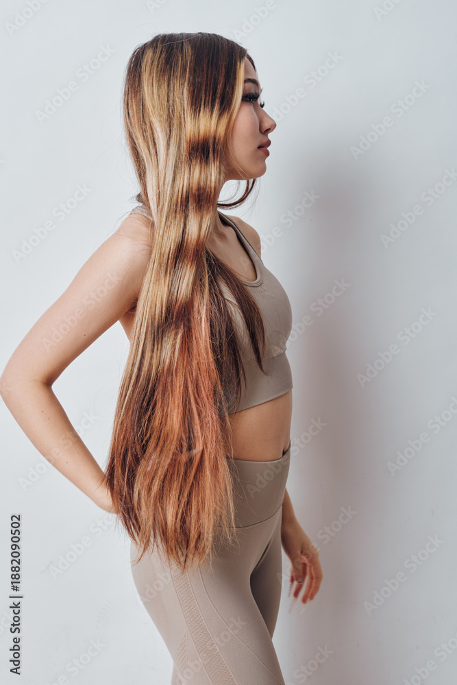 Naklejka premium Young woman with long straight hair standing sideways wearing beige sportswear indoors. Female model with casual pose against plain background showing natural hairstyle and fitness outfit.