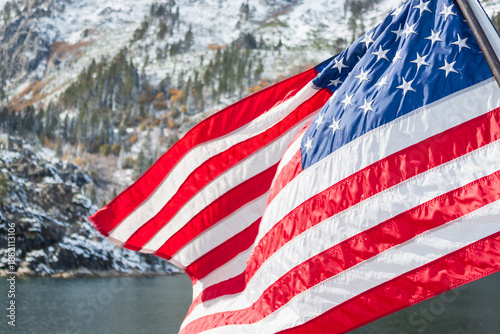 U.S. flag flutters beside steep snow covered rocky cliffs rising from Lake Tahoe.