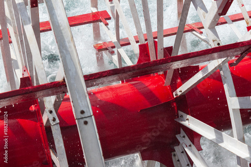 Centered view of a riverboat paddlewheel hub painted red, framed by intersecting silver struts and splashing water for a dynamic industrial composition.