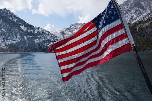 U.S. flag waves from the stern of a boat, trailing ripples across a calm alpine lake surrounded by snowy mountain peaks and soft clouds.