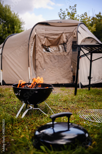 Portable kettle grill with burning charcoal on a grassy campsite, soft-focus tent and camp table behind, sunny outdoor cookout concept with copy space.