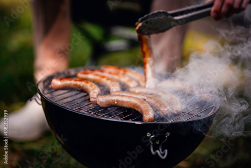 Close-up of hand using tongs to flip sizzling sausages on a smoky charcoal grill during outdoor cooking.