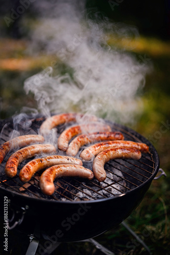 A group of sausages cook on a charcoal grill with smoke rising, set in a grassy outdoor environment.