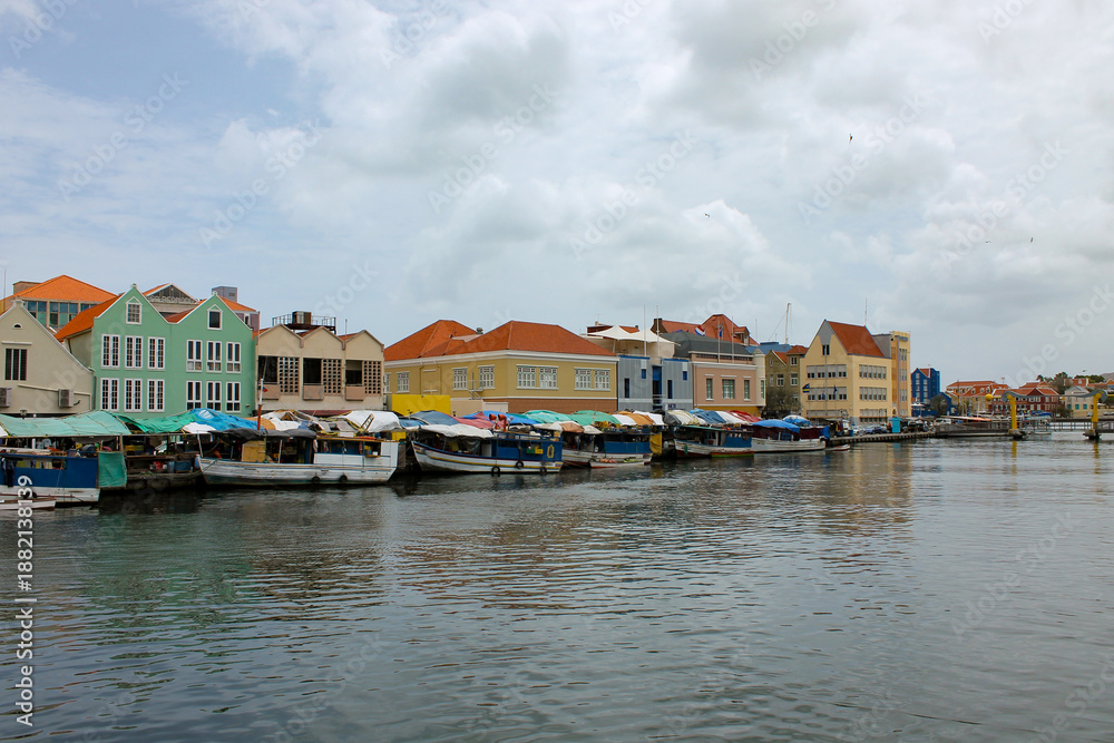 Naklejka premium A vibrant view of the floating market with Venezuelan boats on the water and the historic colonial buildings along the Waaigat in the background in Willemstad, Curaçao. This coastal scene highlights l