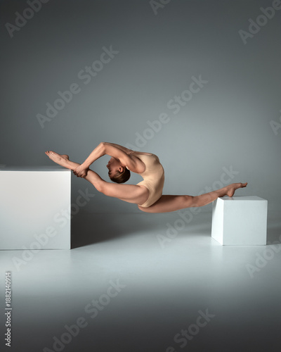 Beautiful young woman limber exerciser in the studio.  Young woman in beige bodysuit posing on cube against blue studio background
