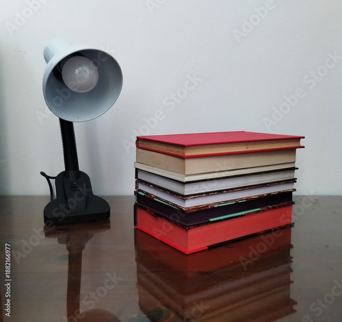 A stack of books on a reflective glass table next to a lamp