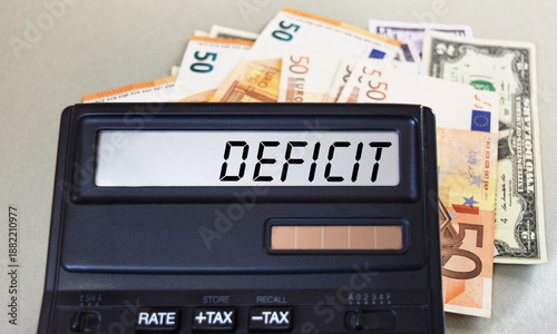 DEFICIT text written on a calculator next to the background of euro banknotes. Finance or business concept.